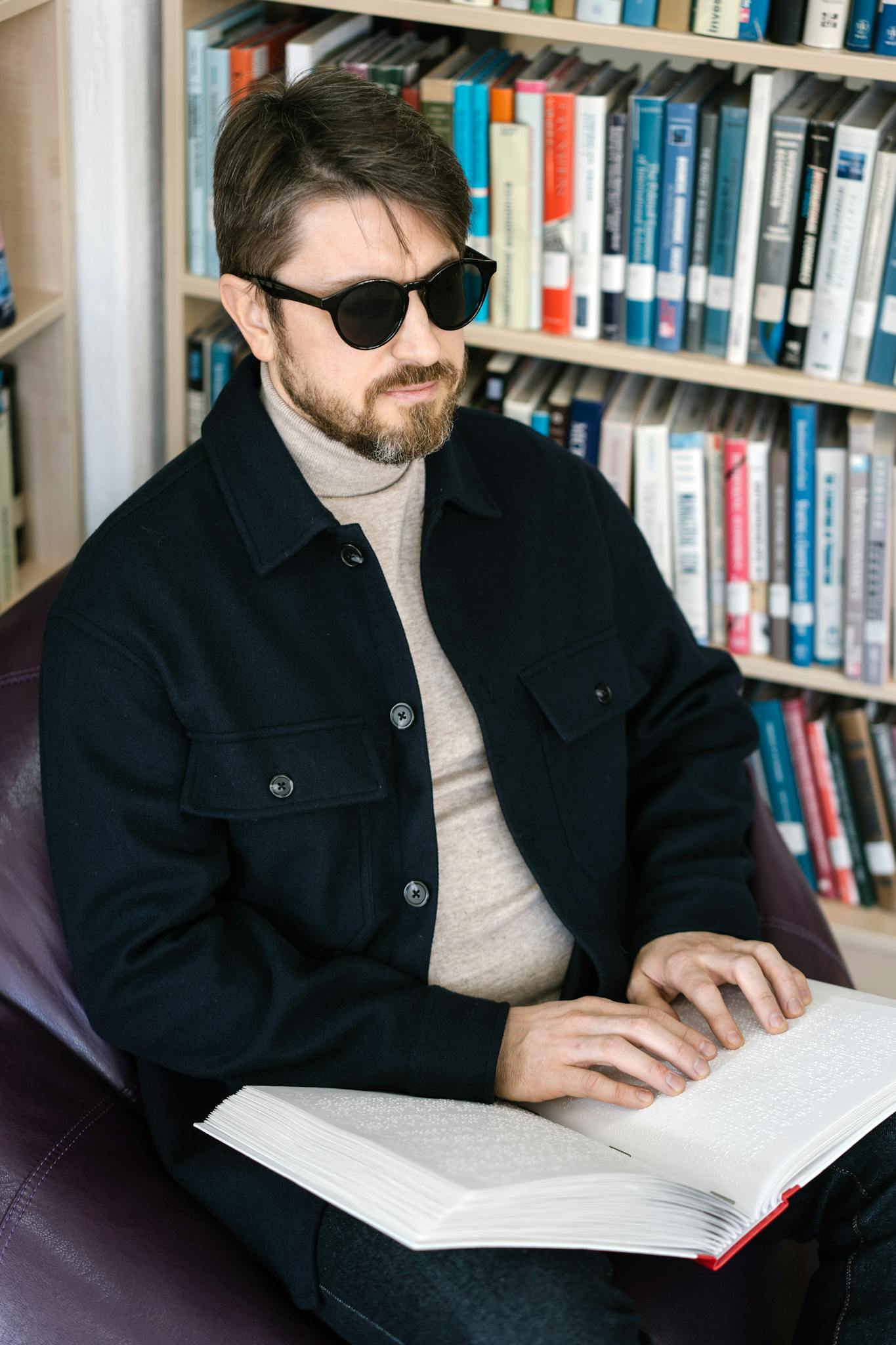 A blind man in sunglasses reads a Braille book in a library, illustrating accessibility.